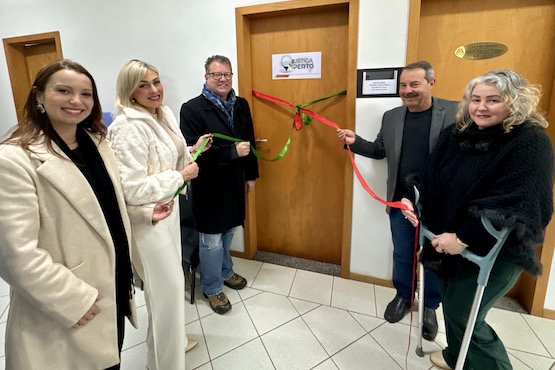 A imagem mostra cinco pessoas em frente a uma porta de madeira, participando de uma cerimônia de inauguração. A porta tem uma fita vermelha e verde amarrada. Da esquerda para a direita: 1. Uma mulher jovem com cabelo castanho claro, usando um casaco bege. 2. Uma mulher loira, usando um casaco branco. 3. Um homem de óculos, usando um casaco escuro e cachecol. 4. Um homem de bigode, usando um blazer escuro e segurando a fita. 5. Uma mulher de cabelo grisalho, usando um casaco preto e segurando a fita e uma muleta.  Na porta, há uma placa com o logotipo e o nome "Justiça + Perto". Ao lado, há outra porta com uma placa dourada. O ambiente parece ser um corredor interno com piso de cerâmica clara.