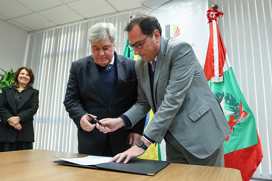 A imagem mostra três pessoas em um ambiente formal, provavelmente um escritório ou sala de reuniões. Duas delas estão em destaque, assinando um documento sobre uma mesa de madeira. À esquerda, um homem de cabelos grisalhos, vestindo um terno escuro, está segurando uma caneta. À direita, outro homem, também de cabelos grisalhos, mas mais curtos, está vestindo um terno cinza e óculos, e parece estar assinando o documento. Ao fundo, uma mulher de cabelos castanhos e vestindo um casaco preto está sorrindo. Atrás deles, há duas bandeiras: uma do Brasil e outra de Santa Catarina. As bandeiras estão posicionadas ao lado de uma parede com persianas verticais brancas.