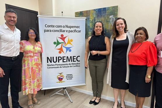 A imagem mostra cinco pessoas posando para uma foto ao lado de um banner. O banner tem o texto "Conte com o Nupemec para conciliar e pacificar" e "NUPEMEC Núcleo Permanente de Métodos Consensuais de Solução de Conflitos". Abaixo do texto, há um logotipo colorido com figuras humanas estilizadas em círculo. Da esquerda para a direita: 1. Um homem de camisa branca e calça preta, sorrindo. 2. Uma mulher de óculos, vestido colorido com predominância de rosa e amarelo. 3. Uma mulher de blusa preta sem mangas e calça verde, sorrindo. 4. Uma mulher de vestido preto e um casaco claro, sorrindo. 5. Uma mulher de blusa vermelha e saia preta, sorrindo. Ao fundo, há uma parede clara com um quadro colorido pendurado. O chão é de madeira clara.