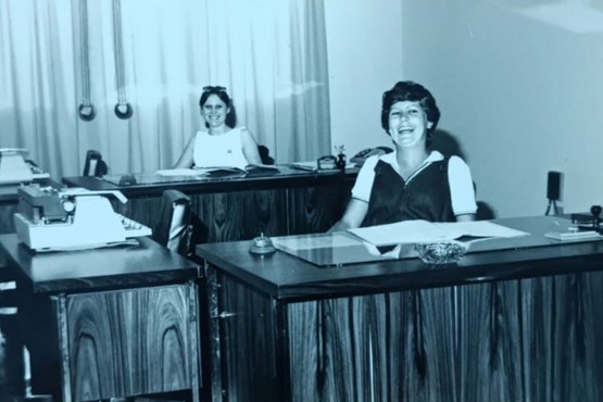 A foto em preto e branco mostra duas mulheres em um escritório antigo. Elas estão sentadas em mesas de madeira com máquinas de escrever. A mulher à direita está mais próxima da câmera e sorri amplamente. Ela usa uma blusa de manga curta e um colete. A mulher ao fundo, à esquerda, também sorri e está sentada em uma mesa similar. Ao fundo, há uma cortina clara e alguns objetos de escritório. O ambiente parece ser bem iluminado.