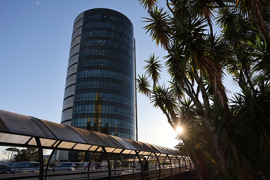 A imagem mostra um edifício alto e cilíndrico com muitas janelas de vidro, refletindo a luz do sol. O prédio está localizado à esquerda da imagem. À direita, há várias palmeiras altas, e o sol está se pondo ou nascendo, criando um efeito de luz entre as folhas das palmeiras. Na parte inferior da imagem, há uma passarela coberta, provavelmente para pedestres, que se estende horizontalmente. Ao fundo, é possível ver alguns carros estacionados.