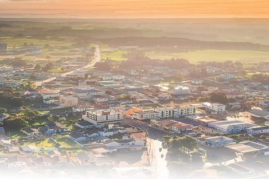 A imagem mostra uma vista aérea de uma cidade ao entardecer. No primeiro plano, há várias casas e edifícios de pequeno e médio porte. Mais ao fundo, é possível ver uma estrada principal que atravessa a cidade, cercada por mais construções e áreas verdes. O horizonte está iluminado por um céu alaranjado, típico do pôr do sol, que dá um tom dourado à paisagem. A imagem transmite uma sensação de tranquilidade e fim de tarde em uma cidade de porte médio.