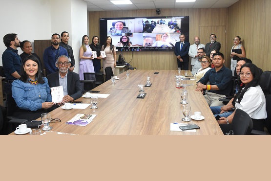 A imagem mostra uma sala de reuniões com uma mesa retangular de madeira no centro. Ao redor da mesa, há várias pessoas sentadas e em pé, totalizando cerca de 19 pessoas. Na parede ao fundo, há uma grande tela de videoconferência com várias janelas mostrando pessoas participando remotamente. Na parte esquerda da mesa, uma mulher de cabelo preto e um homem de barba grisalha estão sentados e segurando documentos. Há copos de água e xícaras de café sobre a mesa. As pessoas estão bem vestidas, sugerindo um ambiente formal ou profissional. A sala tem paredes claras e iluminação de teto.