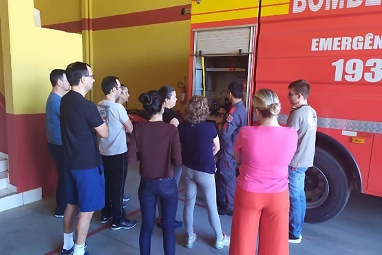 A imagem mostra um grupo de pessoas em pé, aparentemente ouvindo um bombeiro que está explicando algo. Eles estão em frente a um caminhão de bombeiros vermelho, que tem a palavra "Bombeiros" escrita na lateral, junto com o número de emergência "193". O grupo é composto por homens e mulheres de diferentes idades, todos de costas ou de perfil, prestando atenção ao bombeiro. O ambiente parece ser uma garagem ou um quartel de bombeiros, com paredes amarelas e vermelhas.