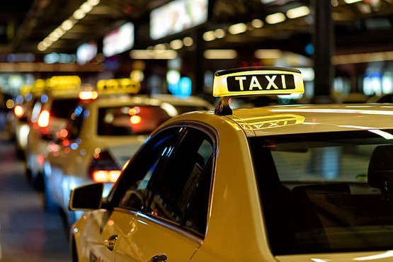 A imagem mostra uma fila de táxis à noite. Os carros estão alinhados um atrás do outro, e cada um tem uma placa luminosa no teto com a palavra "TAXI" escrita em letras maiúsculas. O ambiente parece ser uma área urbana movimentada, possivelmente um aeroporto ou uma estação de trem, devido à presença de várias luzes e sinais ao fundo. As luzes dos táxis e do ambiente criam um efeito brilhante e colorido na imagem.