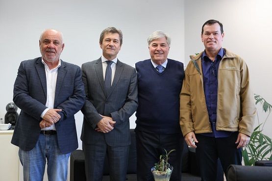 A imagem mostra quatro homens em pé, lado a lado, posando para a foto. Da esquerda para a direita:  1. O primeiro homem é calvo, tem barba e está vestindo um blazer azul escuro, camisa branca e calça jeans. 2. O segundo homem tem cabelo grisalho, está usando um terno cinza escuro com gravata e camisa branca. 3. O terceiro homem também tem cabelo grisalho, está vestindo um suéter azul escuro sobre uma camisa social azul clara. 4. O quarto homem tem cabelo curto e escuro, está usando uma jaqueta bege sobre uma camisa azul escura e calça preta.  Eles estão em um ambiente interno, provavelmente um escritório, com uma planta em um vaso no canto direito da imagem.