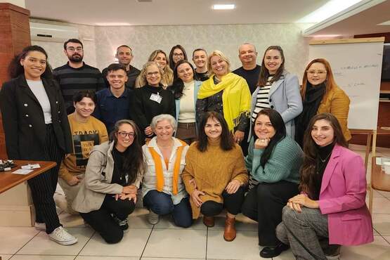A imagem mostra um grupo de pessoas posando para uma foto em um ambiente interno. Há 17 pessoas no total, distribuídas em três fileiras. A maioria está sorrindo e parece estar em um evento ou reunião. Algumas pessoas estão sentadas no chão, outras em cadeiras, e algumas estão em pé. No fundo, há uma parede clara e um quadro branco.