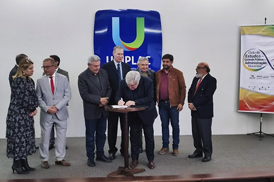 A imagem mostra um grupo de pessoas em um evento formal. No centro, há um homem de cabelos brancos assinando um documento sobre uma mesa redonda pequena. Ao fundo, há um painel com o logotipo da UNIPLAC, que é uma letra "U" estilizada em azul, verde e vermelho. À direita, há um banner com o título "Ciclo de Estudos da Gestão Pública e Administração Municipal". As pessoas ao redor do homem que está assinando o documento estão vestidas formalmente e parecem estar observando a assinatura.