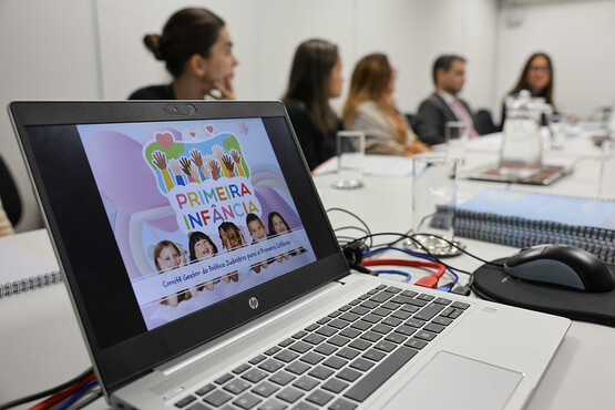 A imagem mostra uma reunião em uma sala de conferências. Em primeiro plano, há um laptop HP aberto sobre a mesa, exibindo uma apresentação com o título "Primeira Infância". A tela do laptop mostra várias mãos coloridas levantadas e fotos de crianças sorrindo. Ao fundo, há seis pessoas sentadas ao redor da mesa, participando da reunião. Elas estão desfocadas. A mesa está organizada com alguns copos de água, uma jarra e alguns documentos.