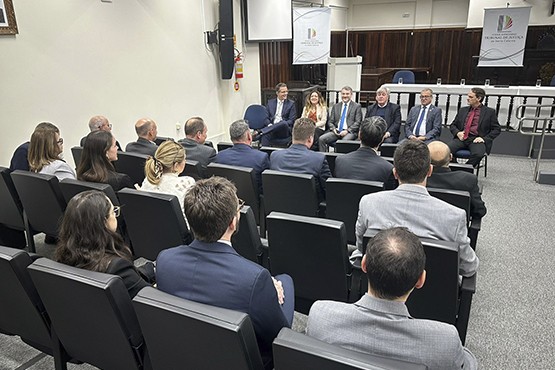 A imagem mostra uma sala de conferências ou reunião, com várias pessoas sentadas em cadeiras dispostas em fileiras. A maioria das pessoas está vestida formalmente, com ternos e roupas sociais. Elas estão voltadas para a frente da sala, onde parece haver uma mesa ou bancada com mais pessoas sentadas, possivelmente conduzindo a reunião. Há também um banner ou cartaz ao lado da mesa, mas o conteúdo não é legível na imagem. A sala tem um ambiente profissional e parece ser um evento formal ou uma reunião de negócios.