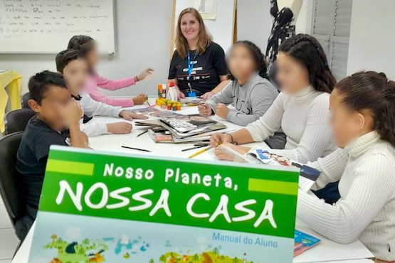 A imagem mostra um grupo de crianças sentadas ao redor de uma mesa, aparentemente em uma atividade educacional. Na mesa, há vários materiais escolares, como canetas coloridas e papéis. Uma mulher, possivelmente a professora ou monitora, está de pé ao fundo, sorrindo. Em primeiro plano, há um cartaz verde com o texto "Nosso Planeta, Nossa Casa". As crianças estão focadas na atividade, e algumas delas estão desenhando ou escrevendo. Ao fundo, há um quadro branco com algumas anotações.