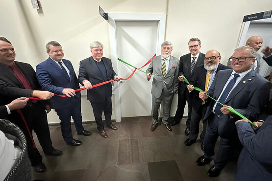 A imagem mostra um grupo de homens de terno participando de uma cerimônia de inauguração. Eles estão segurando uma fita nas cores verde e vermelha, que está prestes a ser cortada. A maioria dos homens está sorrindo e olhando para a câmera. A cerimônia parece estar acontecendo em um ambiente interno, possivelmente um escritório ou uma sala de conferências. Há uma porta branca ao fundo e o chão parece ser de madeira ou um material similar.