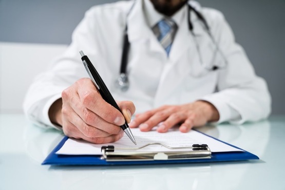 A imagem mostra uma pessoa vestida com um jaleco branco, provavelmente um médico, segurando uma prancheta azul com uma mão e uma caneta com a outra. A pessoa está prestes a escrever ou assinar algo no papel preso à prancheta.