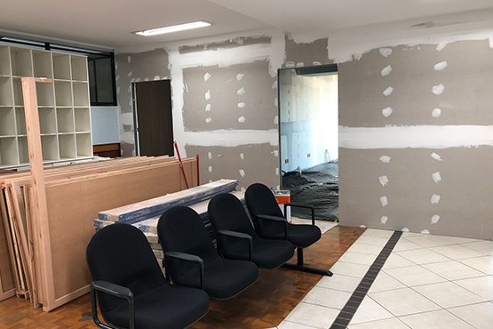 A imagem mostra um ambiente interno em reforma. À esquerda, há uma fileira de cadeiras pretas estofadas, alinhadas uma ao lado da outra. À direita, a parede está em processo de acabamento, com várias marcas de massa corrida, indicando que o drywall está sendo preparado para pintura. No fundo, há uma bancada de madeira clara e, atrás dela, prateleiras vazias. O chão é uma combinação de piso de madeira e cerâmica branca. A iluminação parece ser natural, vinda de uma janela que não está visível na imagem.