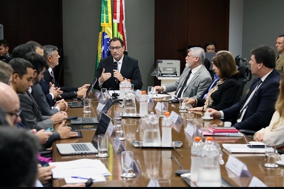 A imagem mostra uma reunião formal com várias pessoas sentadas ao redor de uma mesa retangular. A maioria dos participantes está vestida com trajes formais, como ternos e gravatas. Na mesa, há diversos itens, incluindo laptops, copos de água, garrafas e papéis. Ao fundo, é possível ver bandeiras do Brasil e de outros estados ou instituições. A reunião parece estar ocorrendo em um ambiente oficial, possivelmente governamental ou corporativo.