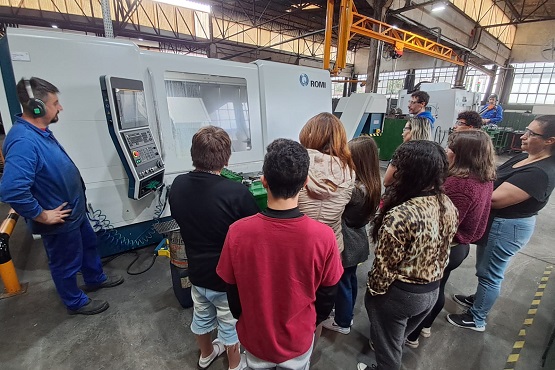 A imagem mostra um grupo de pessoas em uma fábrica ou oficina. Elas estão reunidas em frente a uma máquina grande, possivelmente uma máquina CNC ou algo similar. Um homem de uniforme azul e usando protetores auriculares está operando a máquina ou explicando algo sobre ela para o grupo. As pessoas no grupo estão vestidas de maneira casual e parecem estar prestando atenção na demonstração ou explicação. O ambiente é industrial, com piso de concreto e estruturas metálicas visíveis ao fundo.