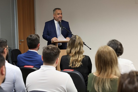 A imagem mostra um homem de terno azul e gravata falando em um púlpito, aparentemente em uma sala de conferências ou auditório. Ele está de frente para uma plateia, que está sentada e prestando atenção. A plateia é composta por várias pessoas, algumas com cabelos loiros e outras com cabelos castanhos.