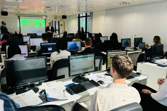 A imagem mostra uma sala de aula ou um ambiente de treinamento com várias pessoas sentadas em frente a computadores. As pessoas estão voltadas para a frente da sala, onde há um projetor exibindo uma apresentação na parede. A sala parece estar cheia, com várias fileiras de mesas e computadores. A maioria das pessoas está concentrada em suas telas ou na apresentação. A imagem está um pouco inclinada, mas é possível ver que o ambiente é bem iluminado e organizado.