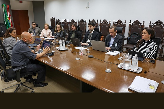 A imagem mostra uma reunião em uma sala de conferências. Várias pessoas estão sentadas ao redor de uma grande mesa de madeira. Entre elas, há homens e mulheres, alguns usando laptops. A mesa está equipada com garrafas de água, copos, um tablet, e outros materiais de escritório.  Ao fundo, pode-se ver uma fileira de cadeiras ornamentadas, sugerindo um ambiente formal, possivelmente pertencente a um tribunal ou órgão governamental. A bandeira do Brasil é visível no canto esquerdo da sala, indicando que o evento está acontecendo no Brasil. Todos parecem estar envolvidos em uma discussão ou apresentação, sugerindo uma visita técnica ou uma reunião de trabalho importante.