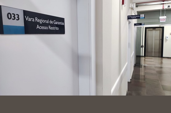A imagem mostra um corredor interno de um prédio. À esquerda, há uma placa na parede com o número "033" e o texto "Vara Regional de Garantias Acesso Restrito". O corredor é iluminado e tem piso de cor escura. No fundo, à direita, há um elevador com portas metálicas. Acima do elevador, há uma placa indicando "Saída". Ao longo do corredor, há outras placas menores indicando diferentes direções ou setores.