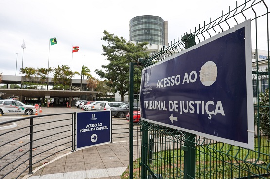 A imagem mostra a entrada de um complexo judicial. Em primeiro plano, há uma placa azul presa a uma grade de metal verde, indicando "Acesso ao Tribunal de Justiça" com uma seta apontando para a esquerda. Ao fundo, há outra placa menor indicando "Fórum da Capital". No lado esquerdo da imagem, há uma área de estacionamento com vários carros estacionados. Ao fundo, há um edifício moderno com uma fachada de vidro e bandeiras do Brasil e de Santa Catarina hasteadas em mastros.