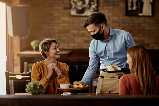 A imagem mostra um garçom servindo duas mulheres em um restaurante. O garçom está usando uma máscara preta e uma camisa azul clara, e ele está segurando uma bandeja com comida. Uma das mulheres, que está de frente para a câmera, tem cabelo curto e veste uma blusa laranja. Ela está sorrindo e olhando para o garçom. A outra mulher, que está de costas para a câmera, tem cabelo comprido e veste uma blusa vermelha. O ambiente do restaurante tem uma decoração rústica, com paredes de tijolos aparentes e móveis de madeira escura. Há também uma pequena planta sobre a mesa.