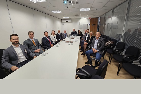 A imagem mostra uma reunião formal em uma sala de conferências. Há várias pessoas sentadas ao redor de uma mesa retangular. A maioria dos participantes está vestida com trajes formais, como ternos e gravatas. A sala é bem iluminada e possui paredes brancas. No fundo, há uma parede de vidro que separa a sala de outra área. No canto inferior esquerdo, há algumas cadeiras vazias.