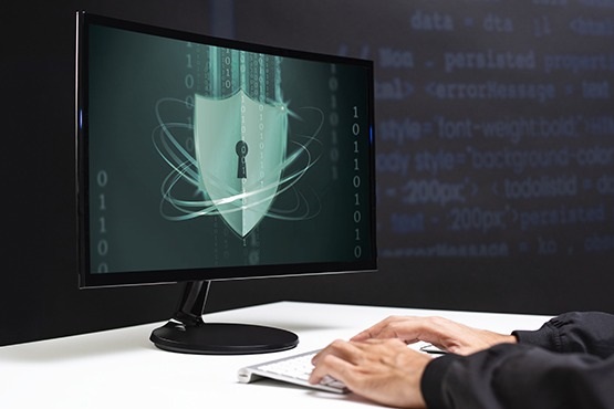 A imagem mostra uma pessoa digitando em um teclado branco, com um monitor de computador à sua frente. Na tela do monitor, há uma imagem de um escudo com um cadeado no centro, simbolizando segurança cibernética. Ao redor do escudo, há linhas e círculos que parecem representar conexões digitais. O fundo da imagem é escuro, com códigos de programação visíveis em azul.