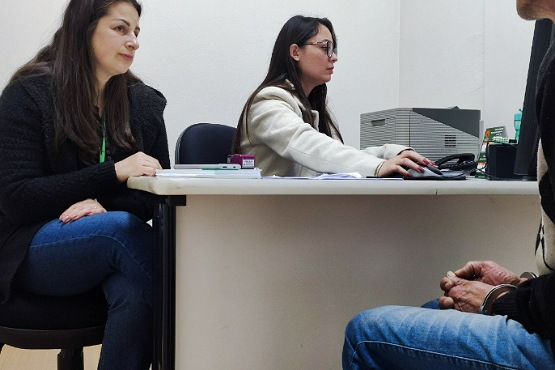 A imagem mostra três pessoas em um ambiente de escritório. Duas mulheres estão sentadas de um lado de uma mesa branca, enquanto um homem está sentado do outro lado. A mulher à esquerda está olhando para o homem e parece estar ouvindo atentamente. Ela tem cabelos longos e escuros e está vestindo um casaco preto e calça jeans. A mulher ao centro está digitando em um teclado de computador e olhando para a tela. Ela tem cabelos longos e escuros, usa óculos e está vestindo um casaco claro. O homem, cuja parte superior do corpo não está visível, está usando uma jaqueta preta e calça jeans. A mesa tem alguns papéis, um carimbo e um computador. O ambiente parece ser uma sala de atendimento.