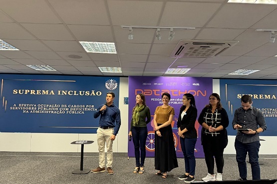A imagem mostra seis pessoas em pé em um palco, participando de um evento. No fundo, há dois painéis azuis com o texto "Suprema Inclusão" e "Efetiva ocupação de cargos por servidores públicos com deficiência na administração pública". Entre esses painéis, há um banner roxo com o texto em destaques e outras informações menores.  As pessoas estão alinhadas lado a lado. Da esquerda para a direita: 1. Um homem de camisa azul e calça clara, segurando um microfone. 2. Uma mulher de blusa verde e calça jeans. 3. Uma mulher de blusa marrom e saia preta. 4. Uma mulher de blusa preta e calça jeans. 5. Uma mulher de blusa preta e calça jeans, segurando um microfone. 6. Um homem de camisa escura e calça preta, olhando para um dispositivo que parece ser um tablet ou celular.  O ambiente parece ser uma sala de conferências com teto de placas brancas e iluminação fluorescente.