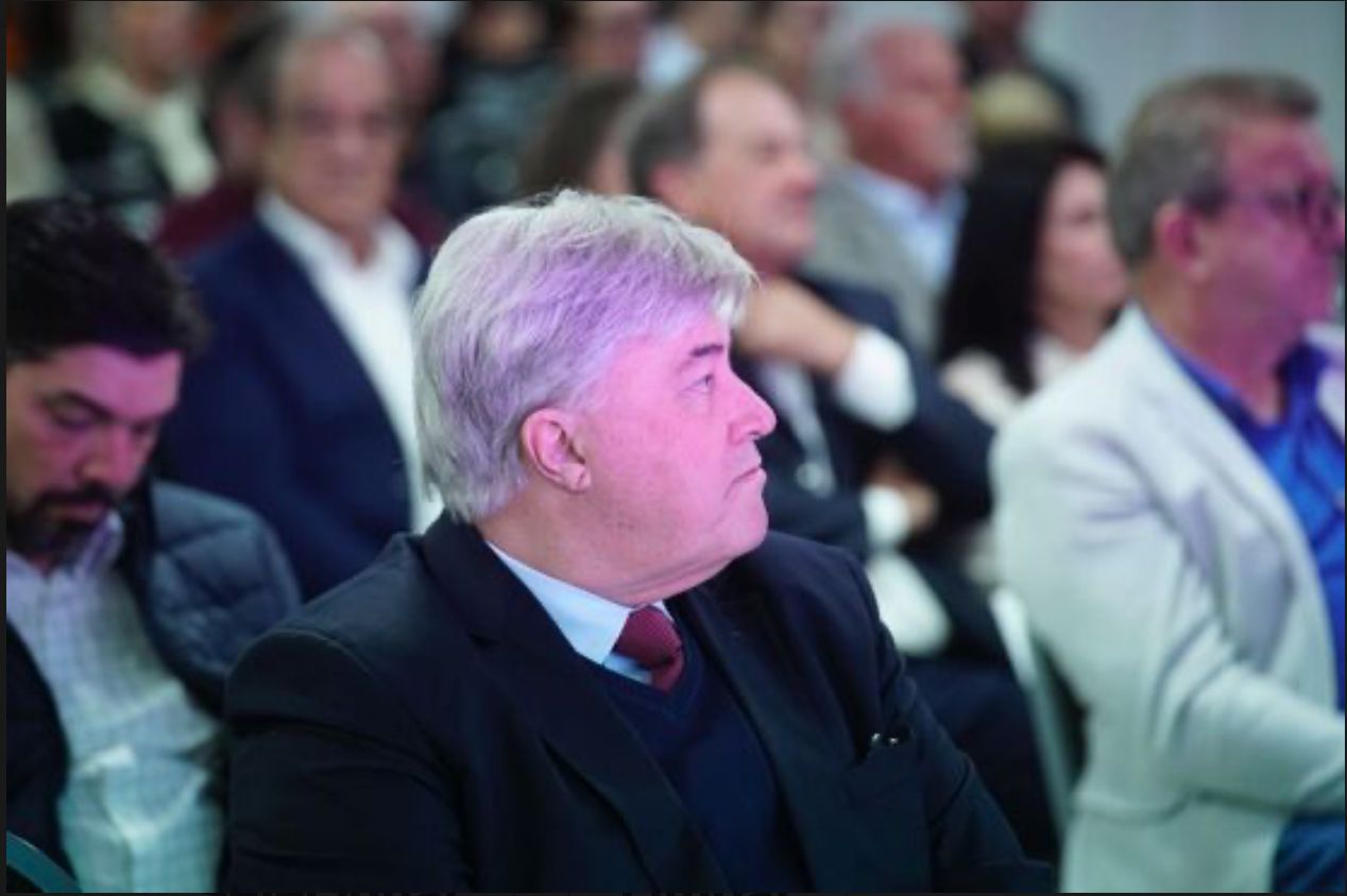 A imagem mostra duas pessoas de perfil, ambas vestindo ternos escuros e gravatas, em frente a uma grande bandeira do Brasil. A bandeira está ao fundo, com suas cores verde, amarelo, azul e branco bem visíveis. A pessoa mais próxima da câmera tem cabelo curto e grisalho, enquanto a pessoa ao fundo tem cabelo castanho.