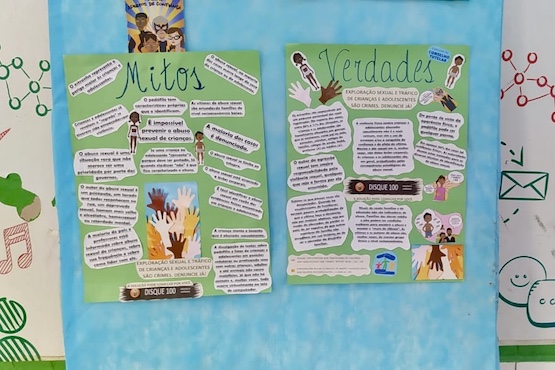 A imagem mostra um mural escolar com dois cartazes principais, um ao lado do outro. O cartaz da esquerda tem o título "Mitos" e o da direita "Verdade". Ambos os cartazes estão sobre um fundo azul claro e possuem textos e ilustrações colados em um fundo verde. No cartaz "Mitos", há várias tiras de papel com textos explicativos e algumas ilustrações, incluindo uma imagem de mãos de diferentes cores se unindo. No cartaz "Verdade", também há várias tiras de papel com textos explicativos e ilustrações, incluindo uma imagem de pessoas conversando. Ao lado esquerdo da imagem, há uma tesoura azul sobre a mesa. Abaixo dos cartazes, há uma parte de uma parede decorada com desenhos e símbolos, incluindo um envelope e algumas conexões em vermelho.