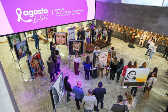 A imagem mostra uma exposição de arte em um espaço fechado. Há várias pessoas observando quadros que estão expostos em suportes. Os quadros parecem ser retratos de pessoas. No canto direito da imagem, há um banner roxo com o texto "Agosto Lilás" e informações sobre o evento, que parece estar relacionado à conscientização sobre a violência contra a mulher. O ambiente está bem iluminado e há uma atmosfera de interesse e interação entre os visitantes.
