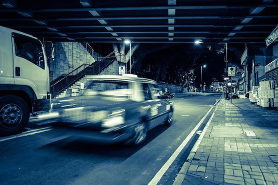 A imagem mostra uma rua à noite com veículos em movimento, capturada com um efeito de desfoque que dá a sensação de velocidade. À esquerda, há uma calçada com ladrilhos e uma faixa amarela pintada no chão. À direita, há um ônibus branco estacionado e uma estrutura de concreto com escadas. A iluminação da rua é fornecida por postes de luz, e ao fundo, é possível ver mais edifícios e luzes.