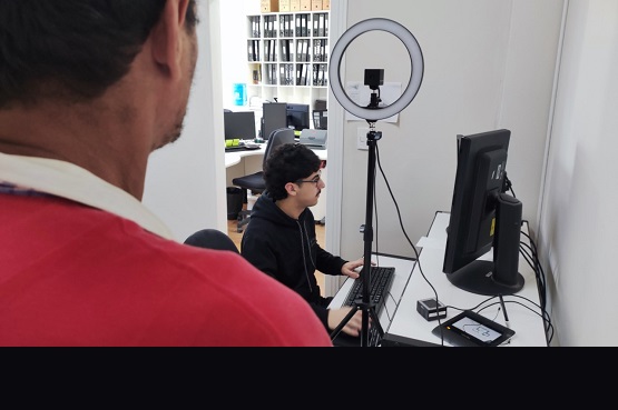 A imagem mostra duas pessoas em um ambiente de escritório. A pessoa em primeiro plano está de costas, vestindo uma camisa vermelha. A segunda pessoa está sentada em frente a um computador, usando um boné preto e uma jaqueta preta. Há um anel de luz montado em um tripé ao lado do computador, provavelmente para melhorar a iluminação. O ambiente parece ser um escritório moderno, com várias mesas e cadeiras ao fundo, além de prateleiras com pastas ou livros.