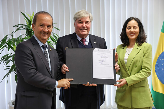 A imagem mostra três pessoas sorrindo e segurando um documento aberto. Da esquerda para a direita, há um homem de terno escuro e gravata, um homem de terno escuro e gravata vermelha, e uma mulher de terno verde claro. Eles estão em um ambiente interno com uma planta ao fundo à esquerda e uma bandeira do Brasil à direita. As cortinas brancas atrás deles deixam a luz entrar, iluminando a cena.