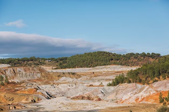 A imagem mostra uma paisagem de mineração a céu aberto. No primeiro plano, há uma área extensa de solo exposto com tons de marrom, laranja e cinza, indicando a extração de minerais. Ao fundo, há uma colina coberta por vegetação verde, principalmente árvores. O céu está claro, com algumas nuvens brancas e uma faixa de nuvens mais escuras no horizonte. A cena transmite uma sensação de exploração e modificação do terreno natural.