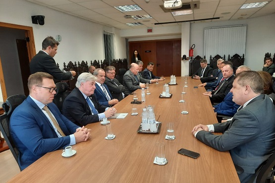 A imagem mostra uma reunião formal em uma sala de conferências. Há uma mesa retangular de madeira no centro, com várias pessoas sentadas ao redor dela. A maioria dos participantes são homens de terno e gravata. Há copos de água e xícaras de café sobre a mesa, além de alguns papéis e um celular. Ao fundo, há uma mulher em pé perto da porta. A sala tem iluminação de teto.
