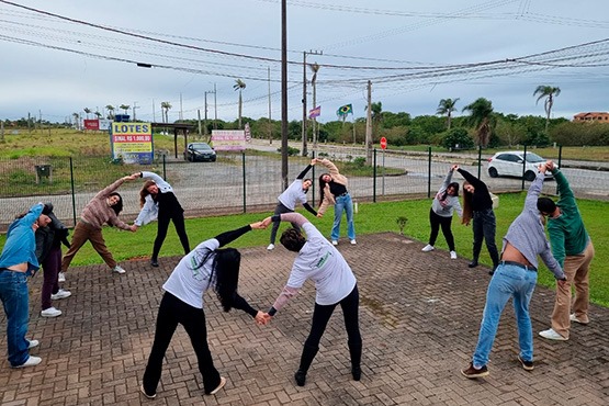 A imagem mostra um grupo de pessoas fazendo exercícios de alongamento ao ar livre. Elas estão em um espaço pavimentado, formando um círculo. Cada pessoa está inclinada para o lado, com um braço estendido acima da cabeça e o outro braço segurando a mão de outra pessoa, formando pares. Ao fundo, há uma cerca de arame e uma estrada com alguns carros passando. Também é possível ver algumas placas e vegetação ao longe. O céu está nublado.