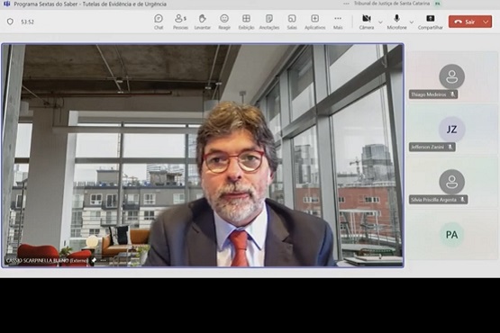 A imagem mostra uma captura de tela de uma videoconferência. No centro da tela, há um homem de meia-idade com cabelo grisalho e barba, usando óculos, terno escuro, camisa branca e gravata vermelha. Ele está sentado em um ambiente que parece ser um escritório moderno, com grandes janelas de vidro ao fundo, através das quais é possível ver prédios e uma paisagem urbana.  Na parte superior da tela, há uma barra de ferramentas com ícones de controle da videoconferência, como microfone, câmera, compartilhamento de tela, entre outros. No canto superior direito, há um botão vermelho de "Sair".  No lado direito da tela, há uma lista de participantes da videoconferência.  Na parte inferior esquerda da imagem, há um texto que diz "Cesar Scarpinella Bueno (Externo)".