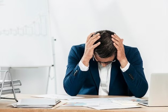 A imagem mostra um homem sentado em uma mesa de escritório, com as mãos na cabeça, parecendo estressado ou preocupado. Ele está vestindo um terno azul e uma camisa branca. Na mesa, há vários papéis espalhados, um laptop e um suporte para documentos. Ao fundo, há um quadro branco com gráficos desenhados.