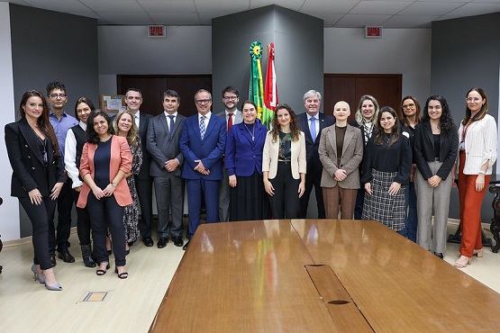 A imagem mostra um grupo de 18 pessoas posando para uma foto em um ambiente de escritório. Eles estão de pé, lado a lado, em frente a uma mesa de madeira grande. No centro da parede ao fundo, há duas bandeiras: a bandeira do Brasil e a de Santa Catarina. As pessoas estão vestidas formalmente, com ternos, blazers e roupas sociais. A maioria está sorrindo para a câmera. O ambiente parece ser uma sala de reuniões.