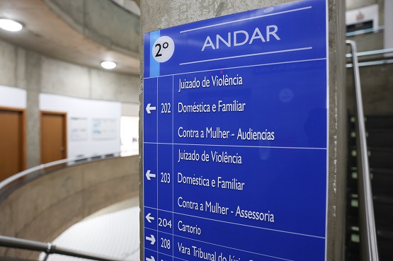 A imagem mostra uma placa azul de sinalização em um prédio, indicando as salas e serviços disponíveis no 2º andar. A placa está fixada em uma coluna de concreto e contém as seguintes informações: 2º ANDAR - Sala 202: Juizado de Violência, Doméstica e Familiar, Contra a Mulher - Audiências (seta para a esquerda) - Sala 203: Juizado de Violência, Doméstica e Familiar, Contra a Mulher - Assessoria (seta para a esquerda) - Sala 204: Cartório (seta para a esquerda) - Sala 208: Vara Tribunal do Júri (seta para a direita) Ao fundo, é possível ver um corredor com portas de madeira e uma escada.