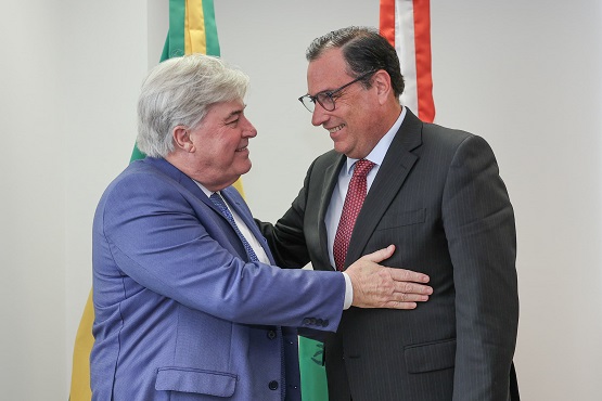 A imagem mostra dois homens se cumprimentando de forma amigável. O homem à esquerda tem cabelo grisalho e veste um terno azul claro com uma camisa branca. Ele está sorrindo e colocando a mão no ombro do outro homem. O homem à direita tem cabelo escuro e usa óculos. Ele veste um terno escuro com uma camisa branca e uma gravata vermelha. Ele também está sorrindo e colocando a mão no braço do homem à esquerda. Ao fundo, há duas bandeiras, uma do Brasil e outra de Santa Catarina.
