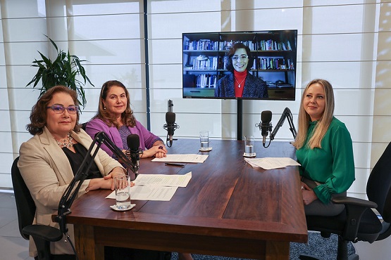 A imagem mostra quatro mulheres em um ambiente de escritório. Três delas estão sentadas ao redor de uma mesa de madeira, enquanto a quarta aparece em uma tela de televisão ao fundo.  Da esquerda para a direita: 1. A primeira mulher está usando um blazer bege e óculos. Ela está sentada à mesa com as mãos cruzadas. 2. A segunda mulher está usando uma blusa roxa e também está sentada à mesa, sorrindo. 3. A terceira mulher está usando uma blusa verde e está sentada à mesa, mais próxima da câmera.  Na tela de televisão ao fundo, há uma quarta mulher, que está usando uma blusa vermelha e está em frente a uma estante de livros.  Na mesa, há microfones e papéis, sugerindo que elas estão participando de uma reunião ou gravação. Ao fundo, há uma planta e janelas com persianas.