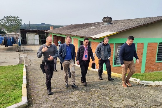 A imagem mostra cinco homens caminhando em uma área externa pavimentada. Eles estão em frente a uma casa de tijolos com janelas e portas verdes. Ao fundo estão algumas árvores e o céu nublado.