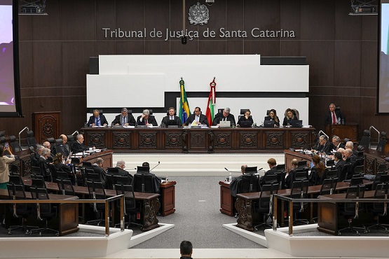 A imagem mostra uma sessão no Tribunal de Justiça de Santa Catarina. A foto está tirada de um ângulo lateral, mostrando várias pessoas sentadas em cadeiras dispostas em formato de "U" ao redor de uma mesa central. Na parte central da imagem, há uma bancada elevada onde estão sentados juízes ou autoridades. Atrás dessa bancada, há bandeiras do Brasil e de Santa Catarina. A parede ao fundo é de madeira escura, e há um brasão no topo com a inscrição "Tribunal de Justiça de Santa Catarina". A imagem parece capturar um momento formal, possivelmente uma audiência ou uma sessão solene.