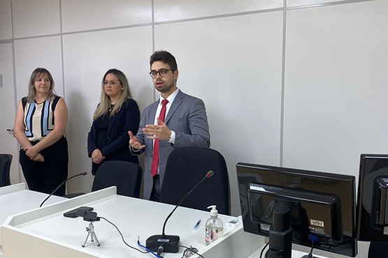 A imagem mostra uma sala de conferências ou tribunal. Há três pessoas em pé atrás de uma mesa comprida na frente da sala, duas mulheres e um homem. Eles parecem estar participando de uma apresentação ou audiência. Na frente deles, há um púlpito com um microfone.