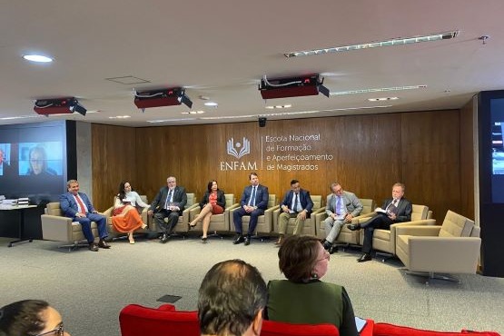 A imagem mostra um painel de discussão em um auditório. Oito pessoas estão sentadas em poltronas dispostas em linha reta no palco. Atrás delas, há uma parede de madeira com o logotipo e o nome "ENFAM" (Escola Nacional de Formação e Aperfeiçoamento de Magistrados). As pessoas estão vestidas formalmente, com ternos e vestidos. No canto esquerdo da imagem, há uma tela de projeção mostrando uma pessoa em videoconferência. Na frente do palco, há algumas pessoas sentadas em cadeiras vermelhas, assistindo ao evento.