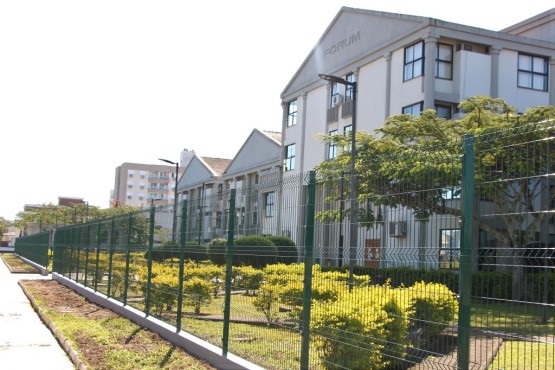 A imagem mostra um prédio de fórum, identificado pela palavra "FÓRUM" escrita na parte superior da fachada. O edifício é de cor clara, com várias janelas e tem um design moderno. Em frente ao prédio, há um jardim bem cuidado com arbustos e árvores. Uma cerca de metal verde delimita a área do jardim. Ao fundo, é possível ver outros edifícios residenciais ou comerciais. O céu está claro, indicando que é um dia ensolarado.